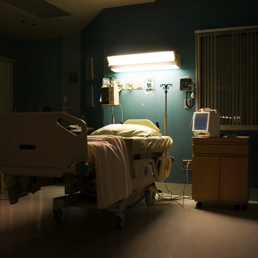 An empty hospital bed in a dark room. The bed is illuminated by a single overhead light