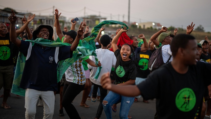 A group of young South African men and women in black and green clothing dance joyously in the street.