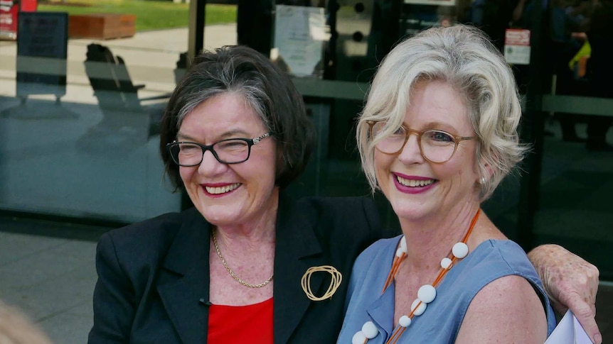 Cathy McGowan, in a red shirt and black blazer, embraces Helen Haines, in a blue blouse. Both are smiling