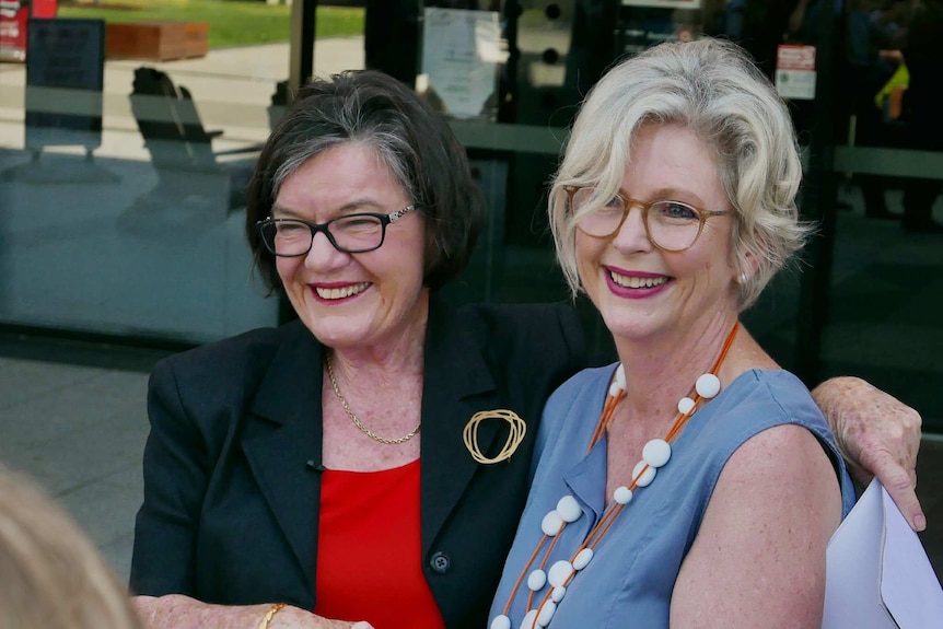 Cathy McGowan, in a red shirt and black blazer, embraces Helen Haines, in a blue blouse. Both are smiling