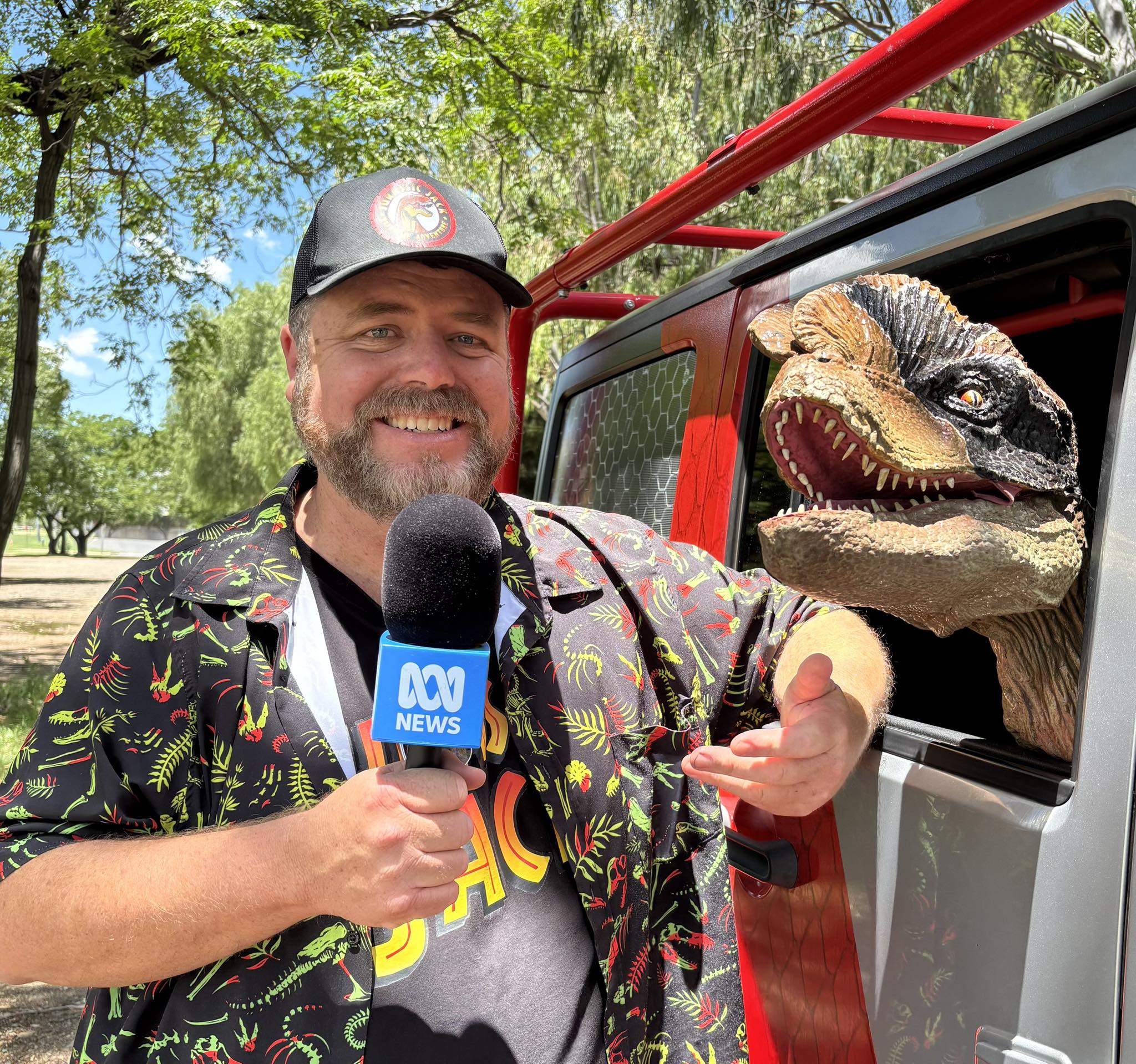 Un hombre con una camisa colorida sostiene un micrófono ABC, su brazo se apoya en la ventanilla de un automóvil por donde asoma la cabeza un modelo de dinosaurio