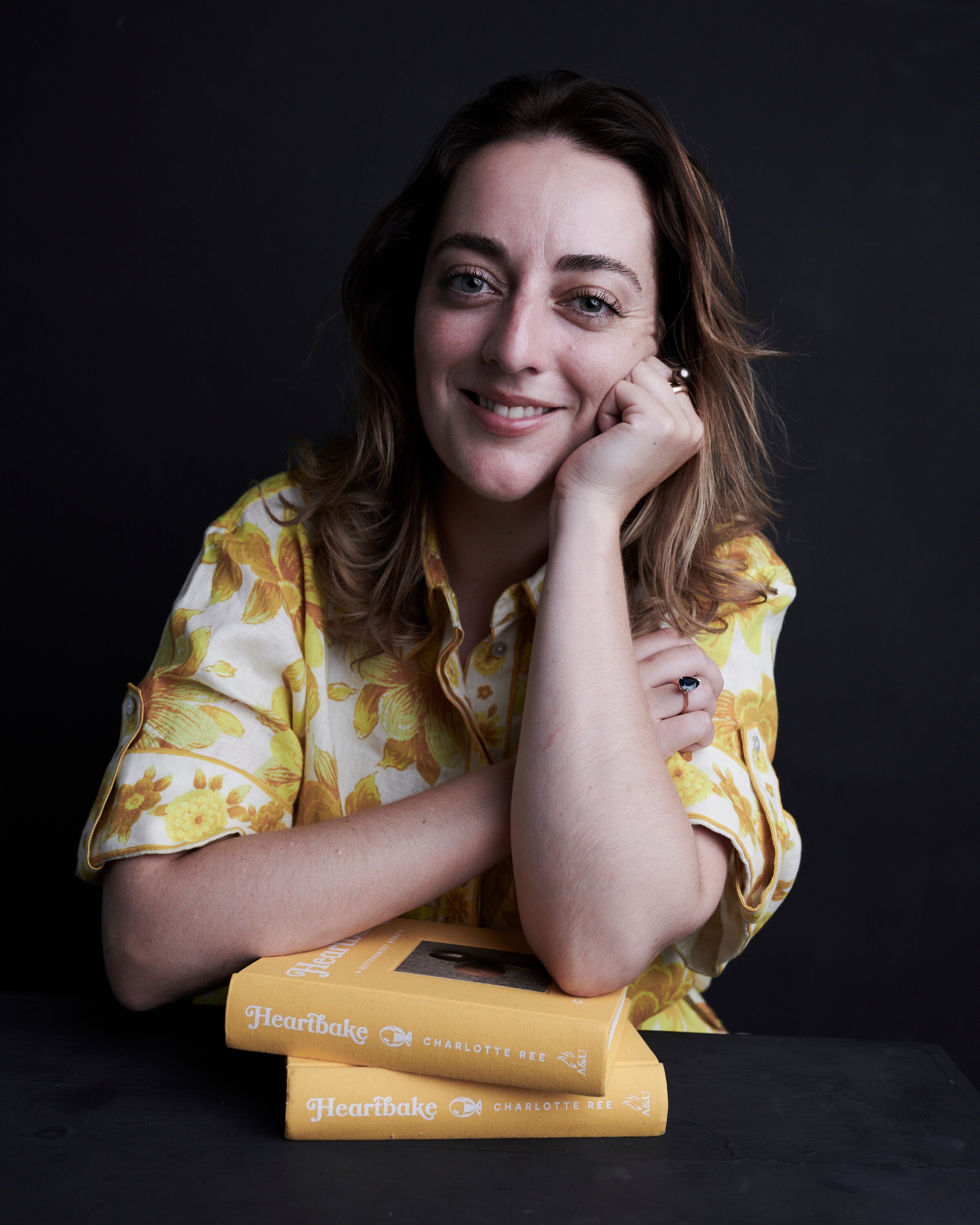 A dark-haired young woman wearing a shirt with a yellow floral pattern with her chin resting on her hand