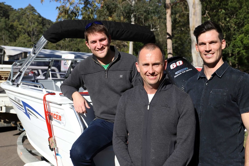 Three man standing in front of a boat in a car park.