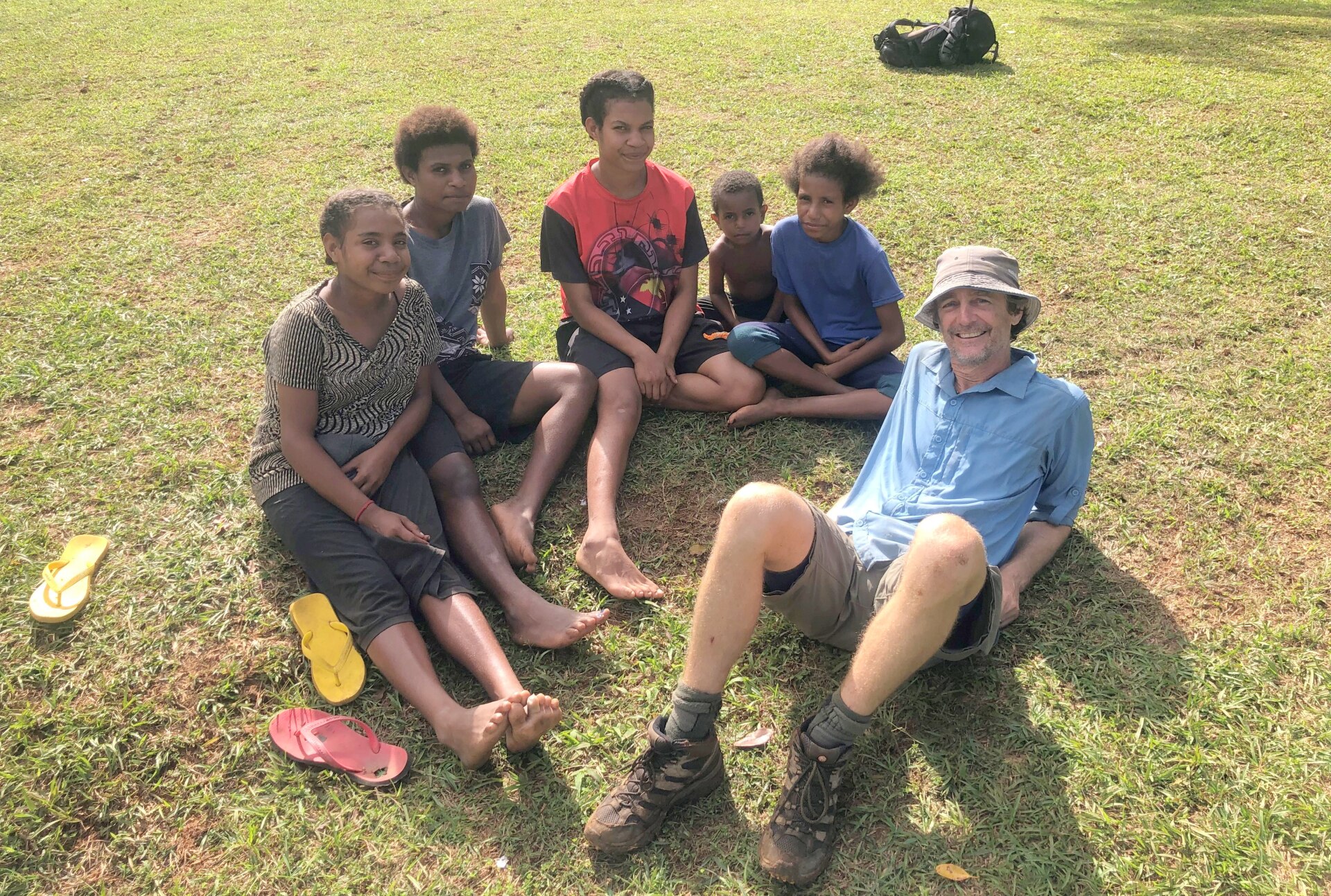Australian man reclining on ground on right, with five Papua New Guinean teenagers and children sitting with him.