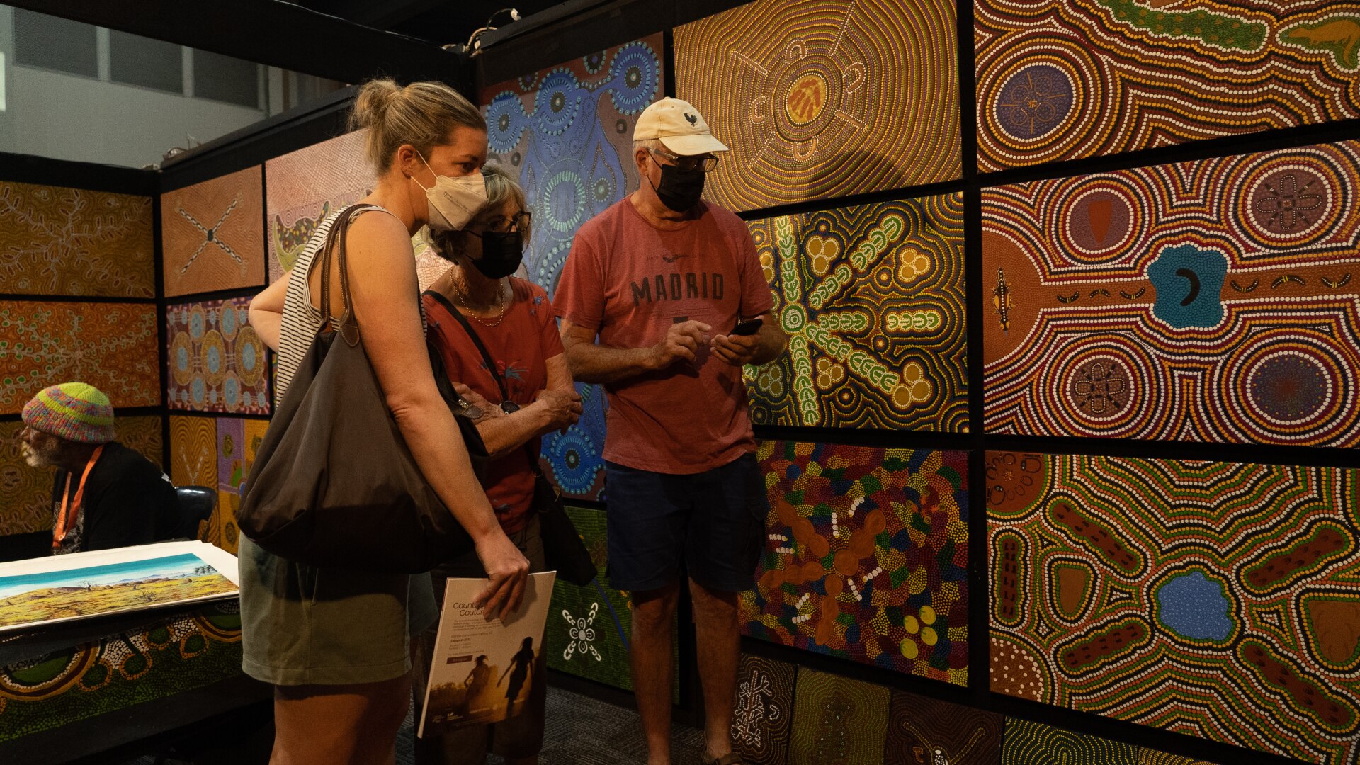 Three people looking at brightly colored traditional Aboriginal paintings hung on the walls of an art gallery.