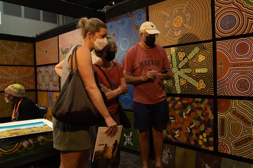Three people looking at brightly colored traditional Aboriginal paintings hung on the walls of an art gallery.