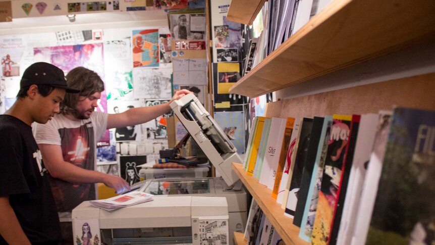 Two men use a photocopier, photocopied magazines on shelves