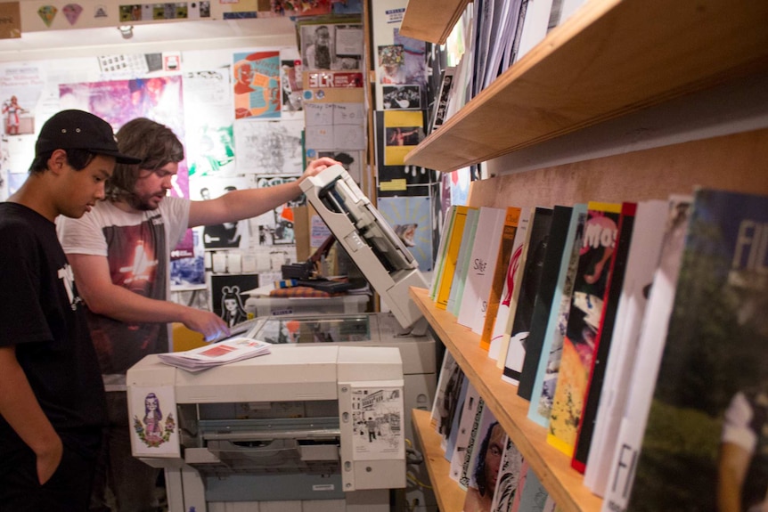 Two men use a photocopier, photocopied magazines on shelves