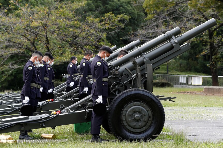 Uniformed personnel bow their head for a moment of silence as they stand behind cannons.