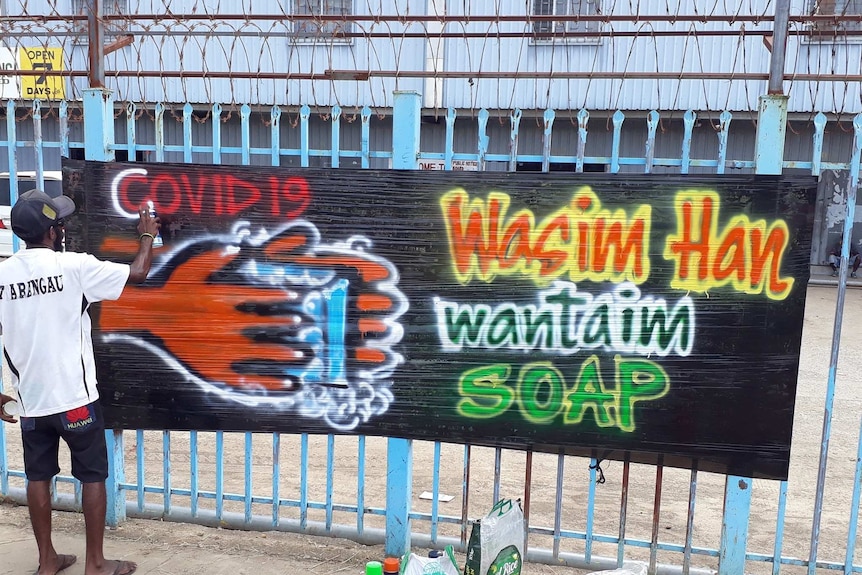 A man wearing a white shirt and black cap spray-paints the words COVID-19 on some cardboard on a fence.