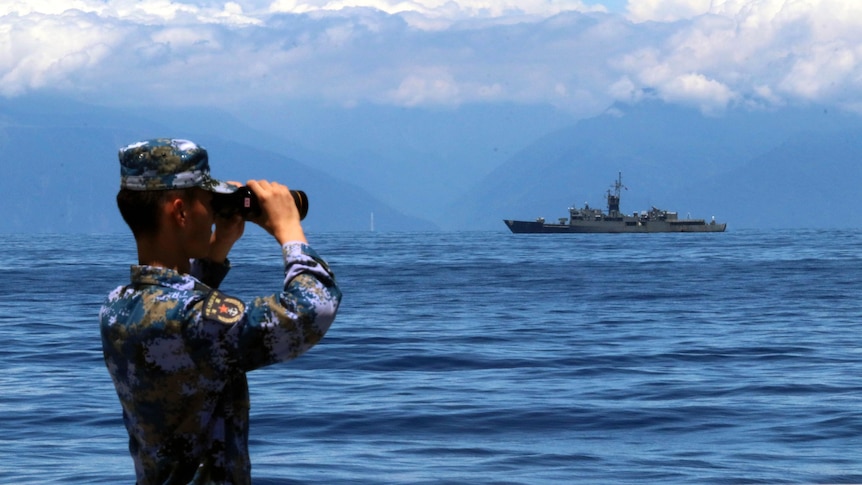 A man in army colours looks through binoculars out to sea where there is a ship in the background. 