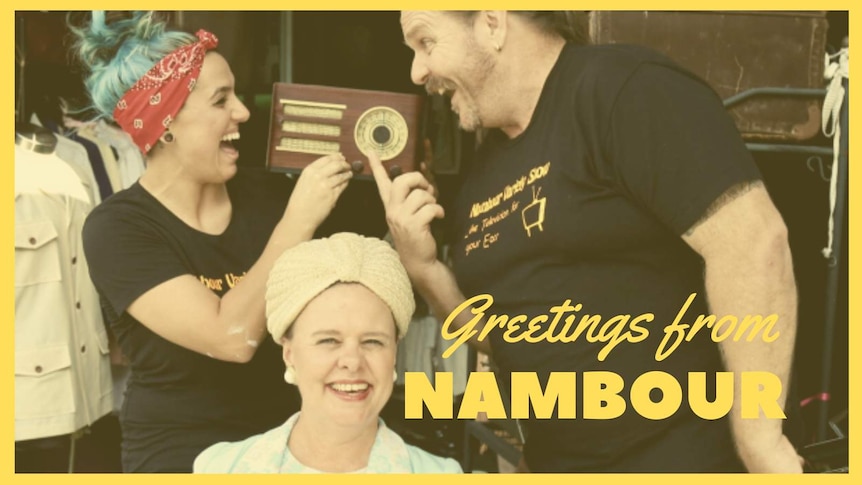 An older woman dressed in 1950s clothing is seated in front of a younger man and woman who are holding an old fashioned radio.