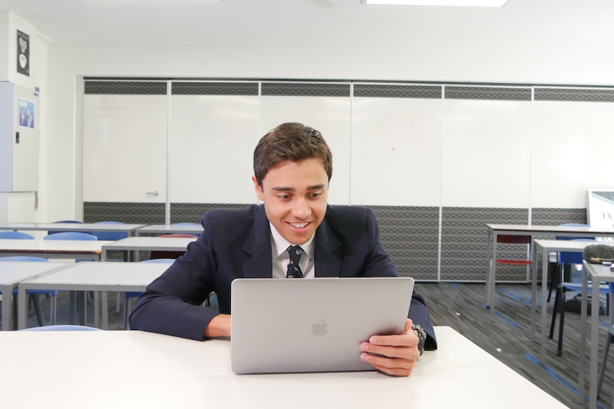 A young man looks at a computer screen. He is in a navy blazer and in a classroom.