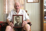 An elderly man sits in a chair holding a framed photo of his younger self on his knees.