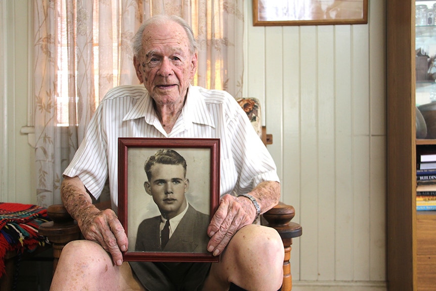 An elderly man sits in a chair holding a framed photo of his younger self on his knees.