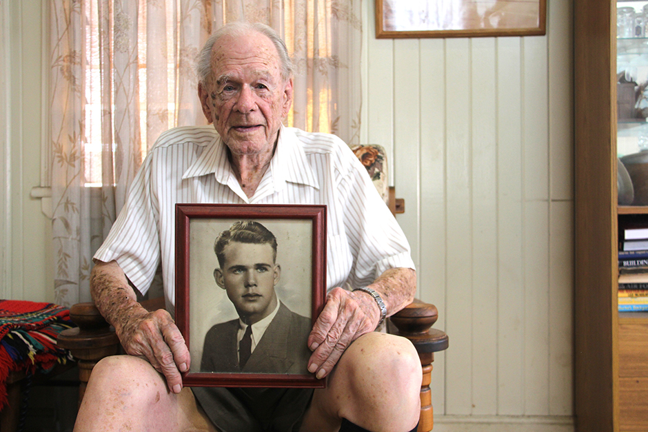 An elderly man sits in a chair with a framed photo of his younger self resting on his knees.