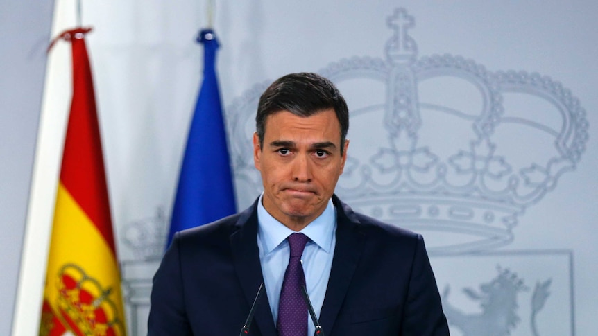 In front of a grey backdrop with a crown, Sanchez looks forlorn at cameras while speaking on a lectern with Spanish and EU flags