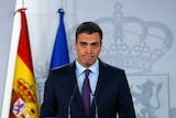 In front of a grey backdrop with a crown, Sanchez looks forlorn at cameras while speaking on a lectern with Spanish and EU flags