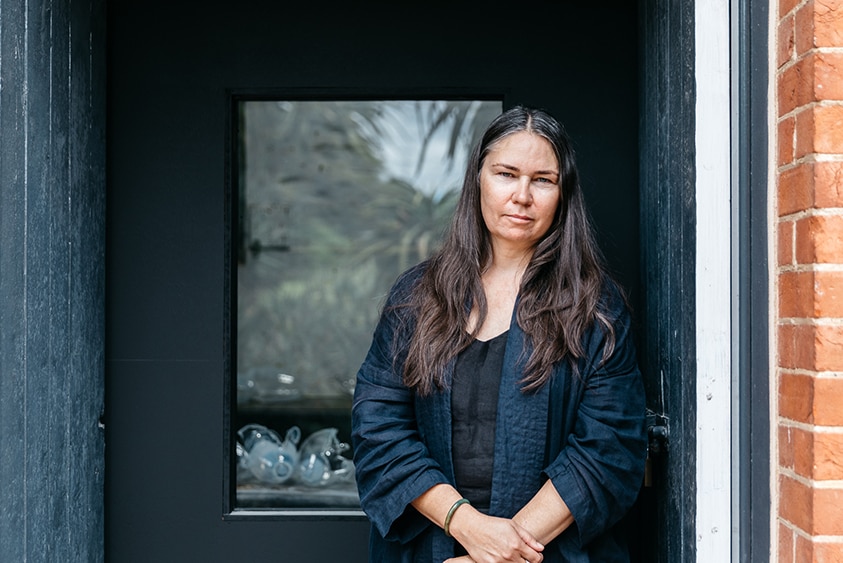 A woman with long dark hair and serious expression stands in wooden doorway of brick building.