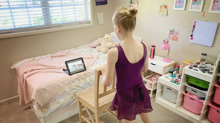 A young girl uses a chair as a ballet barre to follow an online class screened on an iPad in her bedroom.