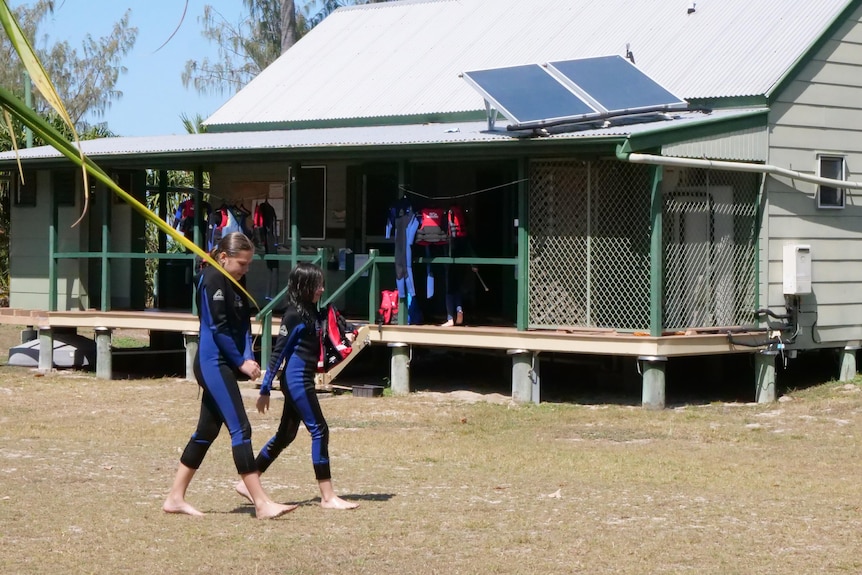Two kids wearing wetsuits walk in front of a hut with a solar panel on the roof.
