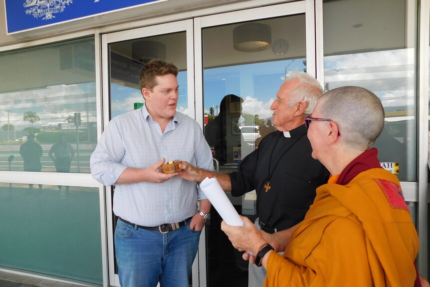 A man standing outside a politician's office greets a priest and a Buddhist nun.