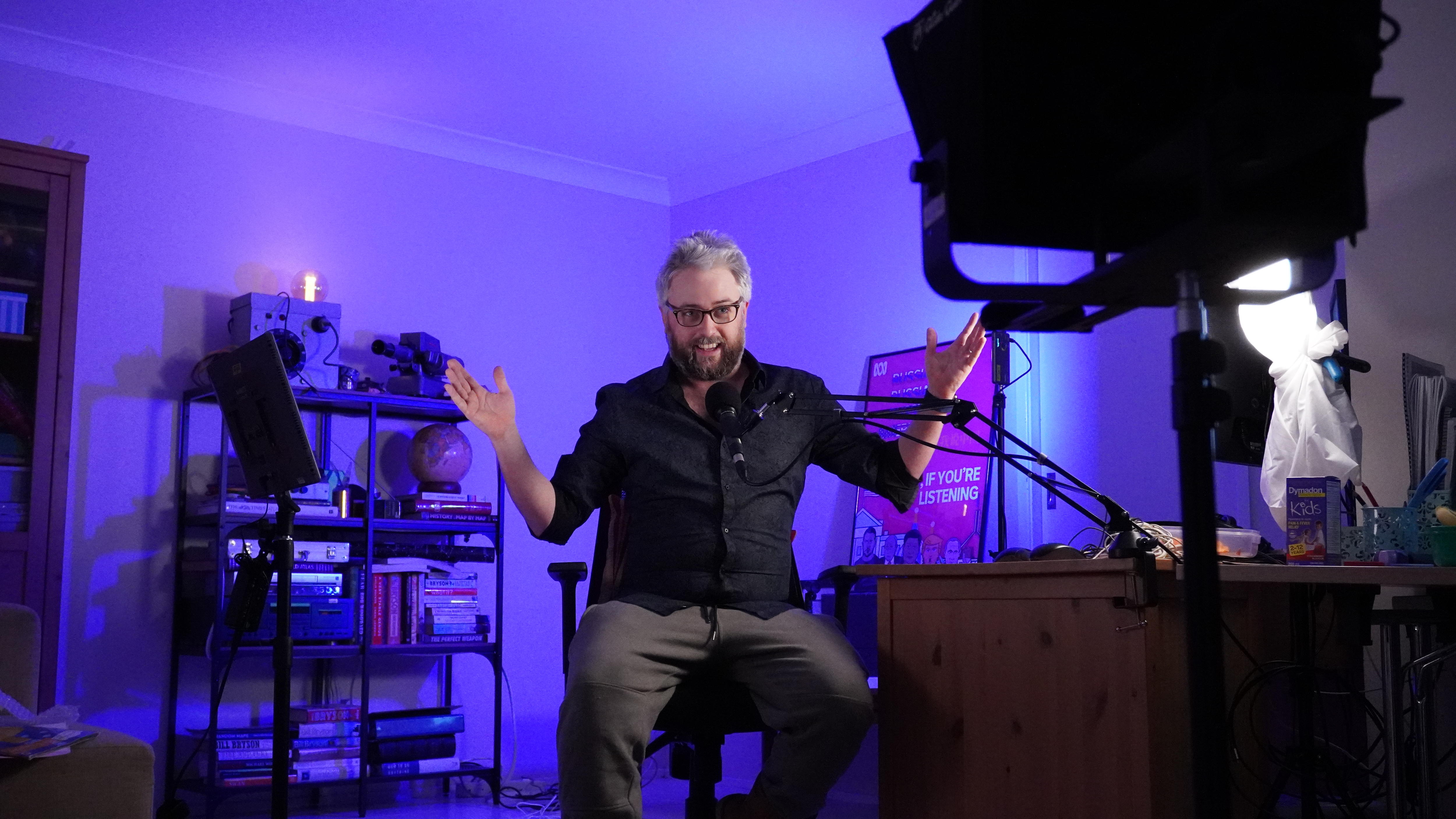 Man sitting on chair in an office with purple lighting, hands apart, talking into a microphone and looking towards a camera. 