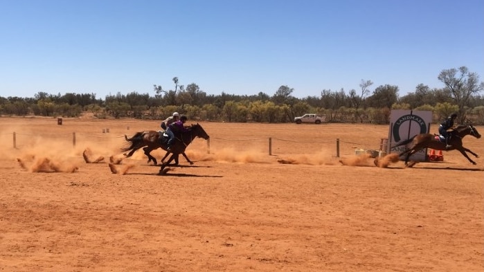 Three horses race on the red dirt track they're approaching the finish line, dust is flying up.