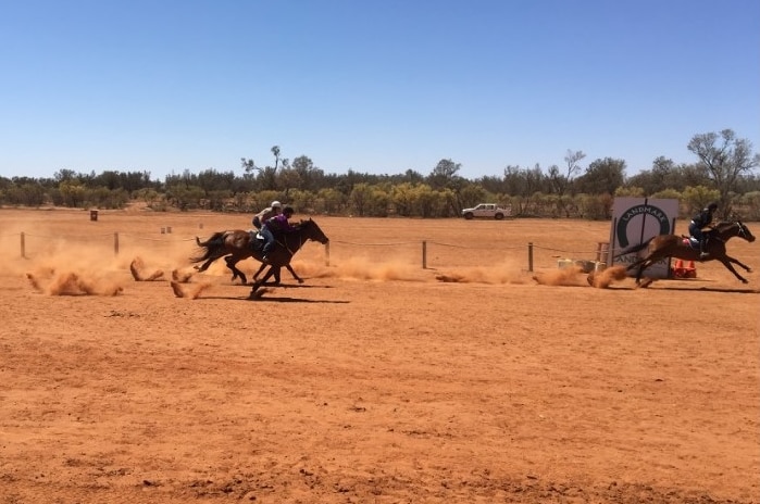 Three horses race on the red dirt track they're approaching the finish line, dust is flying up.