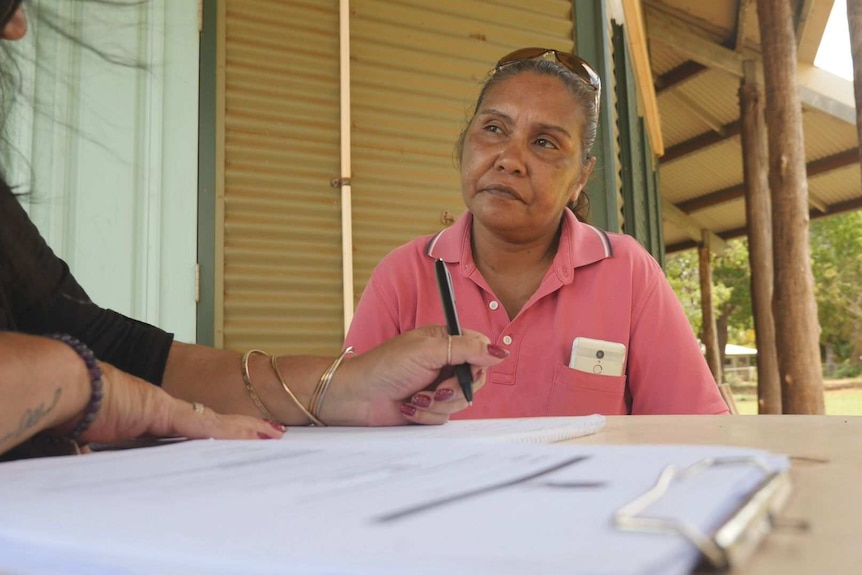 A woman wearing a pink polo shirt looks at another person writing on paper near a clipboard.