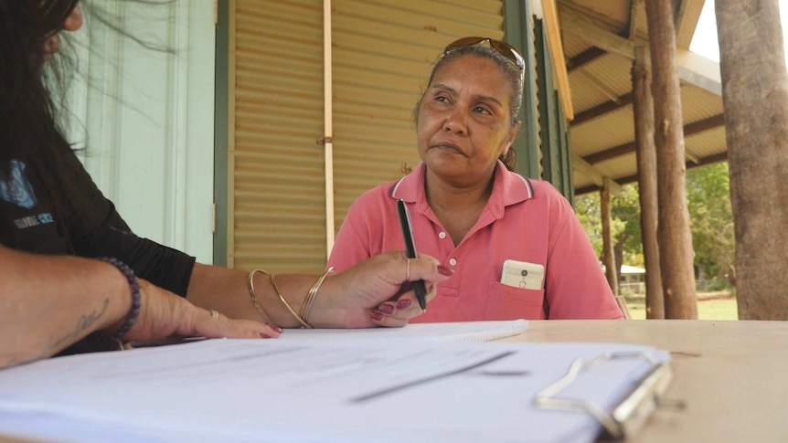 A woman wearing a pink polo shirt looks at another person writing on paper near a clipboard.