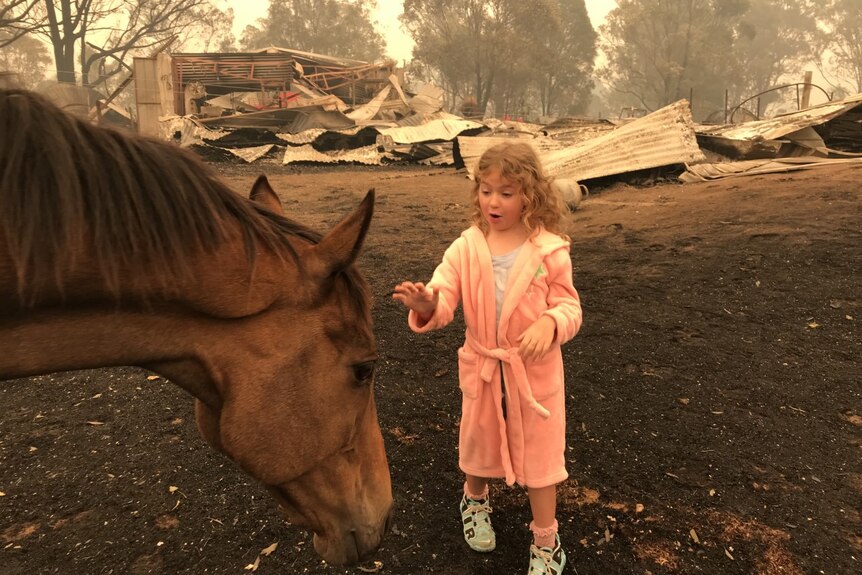 Eight year old child stands in nightgown patting a horse among blackened landscape.