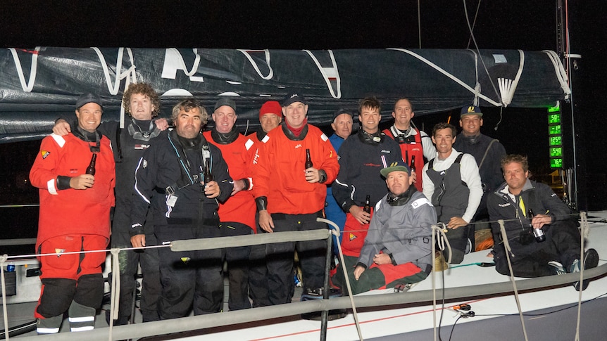 Thirteen men in red, black and crew wet weather coats, grouped together, holding beers on yacht deck at night.