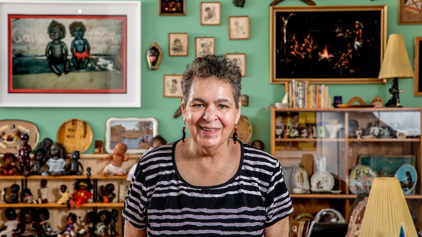 A woman wearing a striped-shirt smiles. Aboriginal kitsch dolls and images fill the background.
