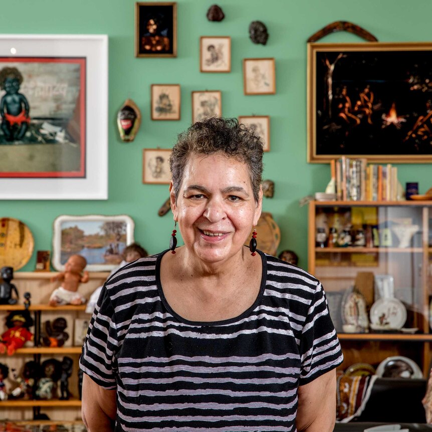 A woman wearing a striped-shirt smiles. Aboriginal kitsch dolls and images fill the background.