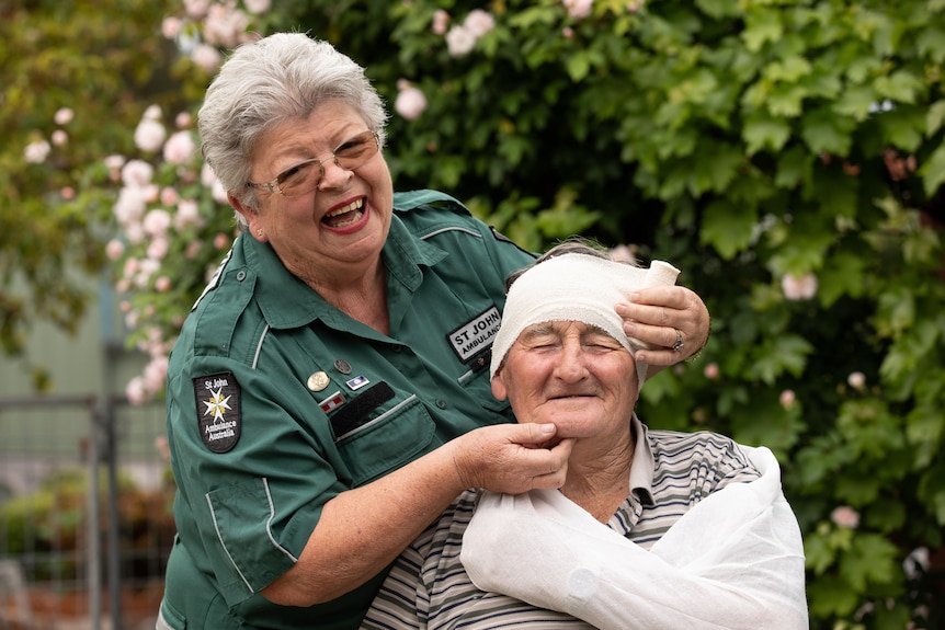 Val laughs while wrapping a man's head in a bandage.