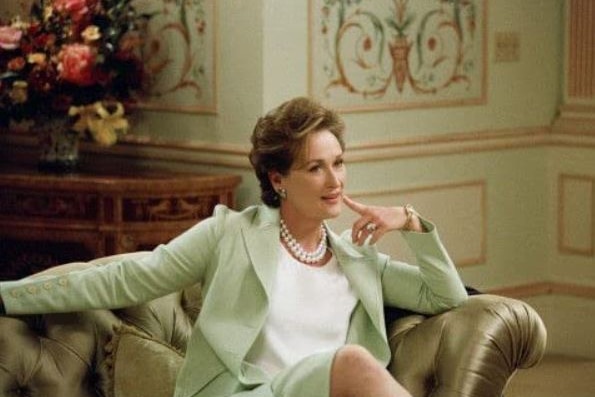 Meryl Streep sits on a chair with her finger pointed at her chin, thinking and smiling.