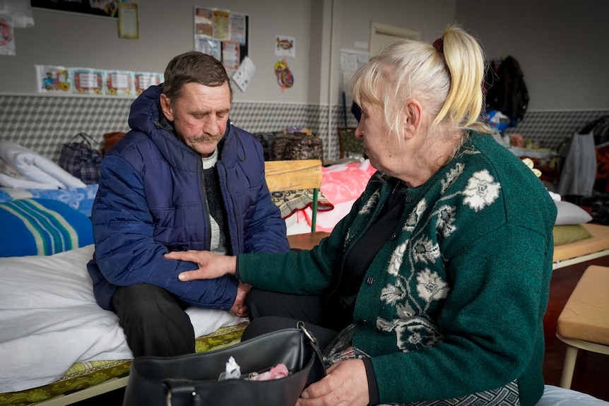 A blonde woman tenderly touches a man's forearm as they sit in a dorm room