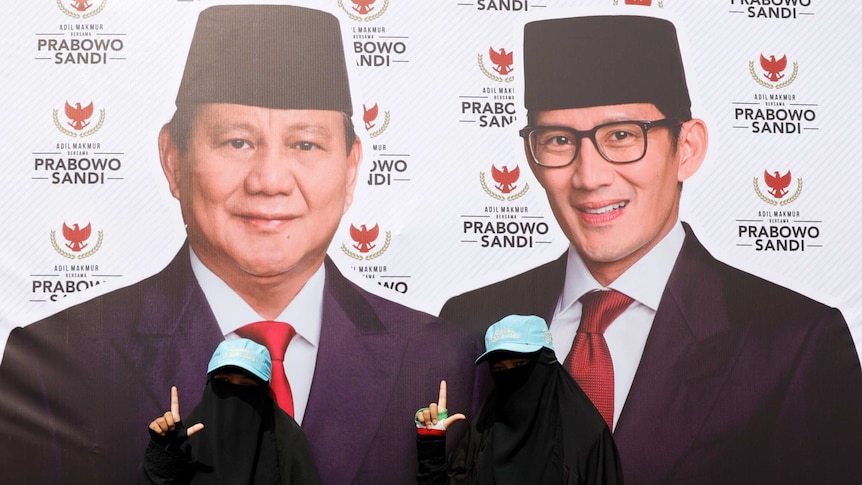 Two women dressed in black, full face veils stand in front of a large political banner in Indonesia.