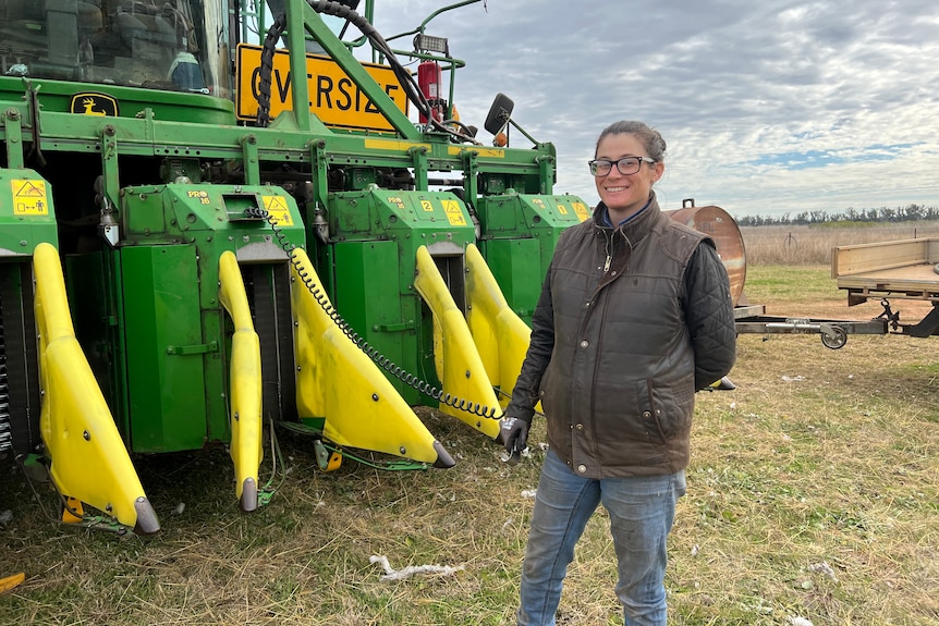 A pregnant woman wearing a sleeveless puffer jacket stands in front of a large green and yellow tractor