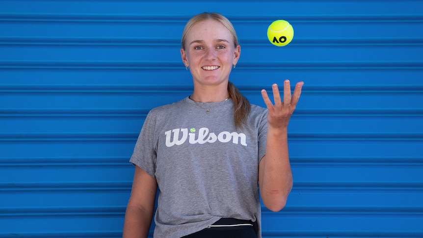 Taylah Preston stands in front of a blue wall, she wears a grey shirt and is throwing a tennis ball in the air, smiling.