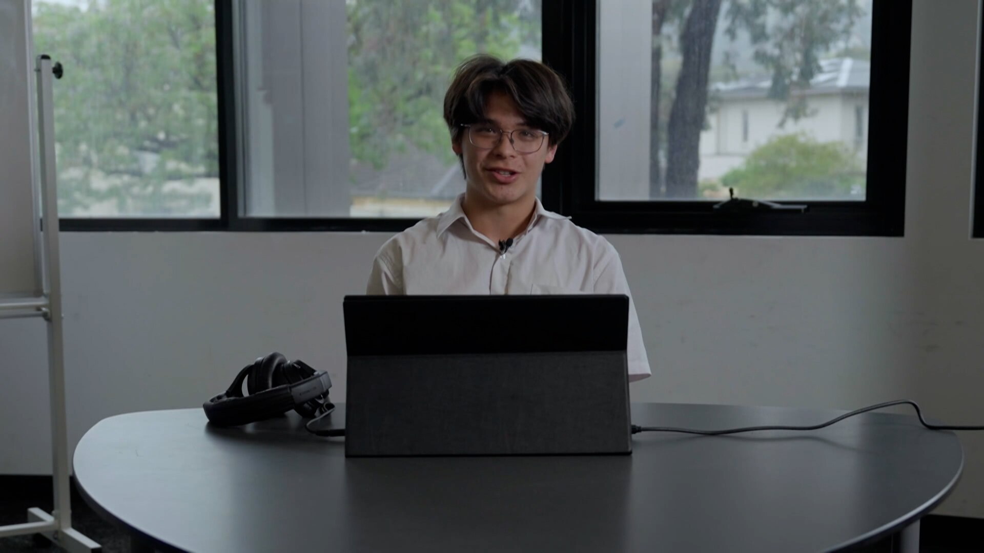 A young man with brown hair and glasses sits at a desk with a computer. He wears a white shirt.