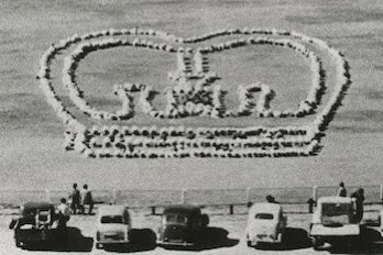 an archival black and white photo of an aerial view of children sitting in the shape of the queen's crown on an oval