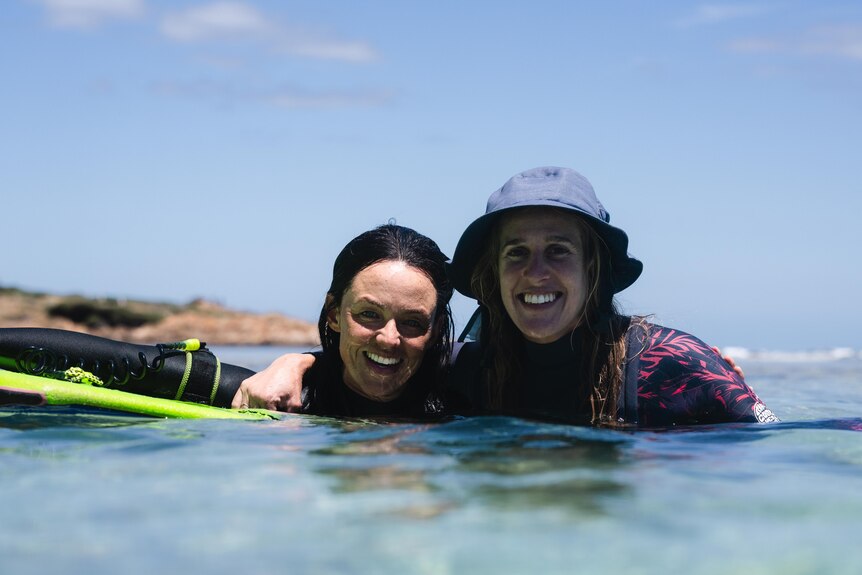 Two women floating on surfboards in the water, embrace and smile at the camera. 