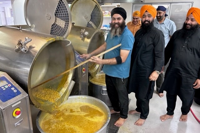 Sikh men wearing turbans gather around a large pot of dahl or curry in an industrial style kitchen.