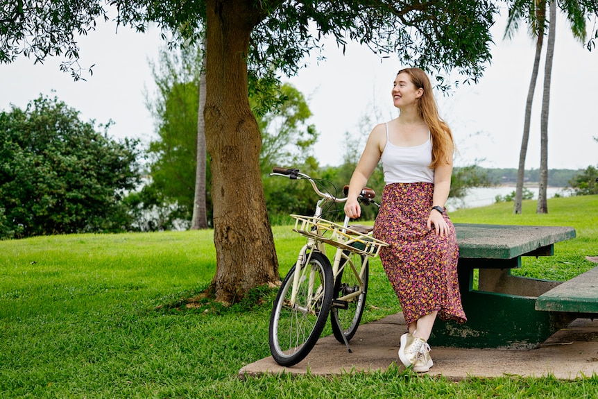 A woman stands outside next to a bike and smiles.
