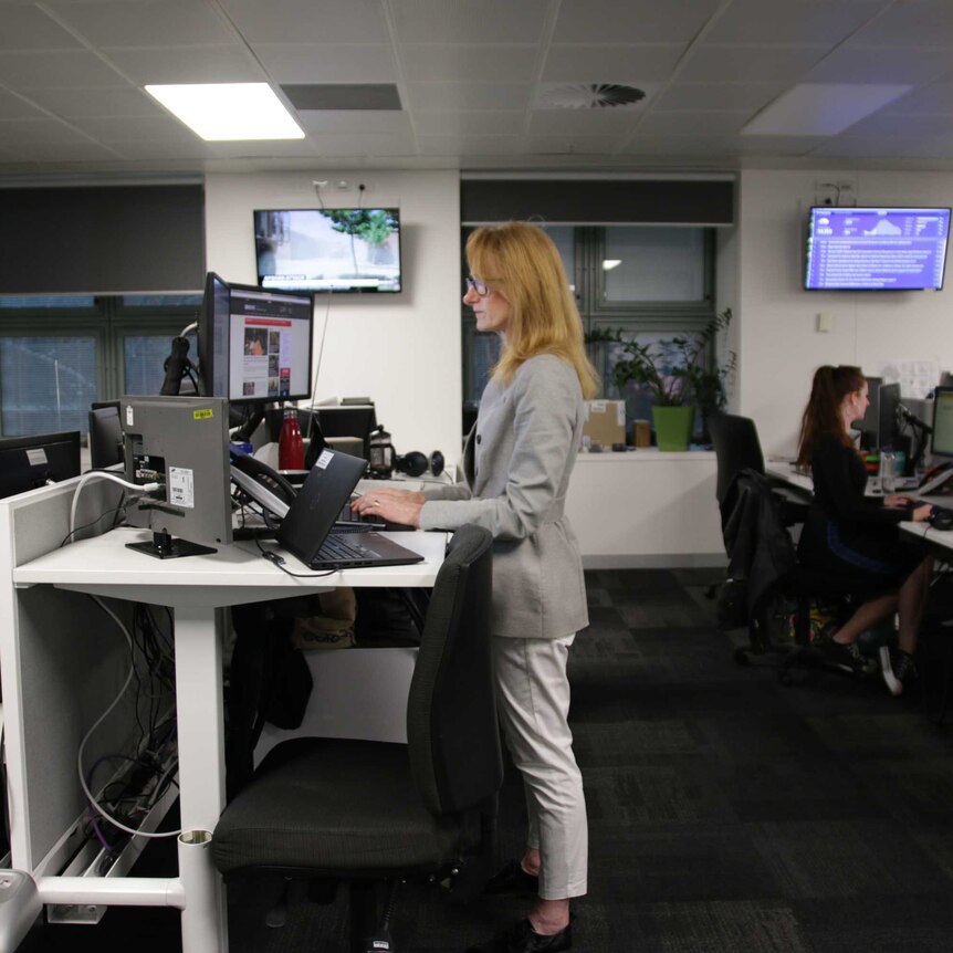 A woman is standing at a desk in an office, another woman is sitting at a desk behind her and out of focus