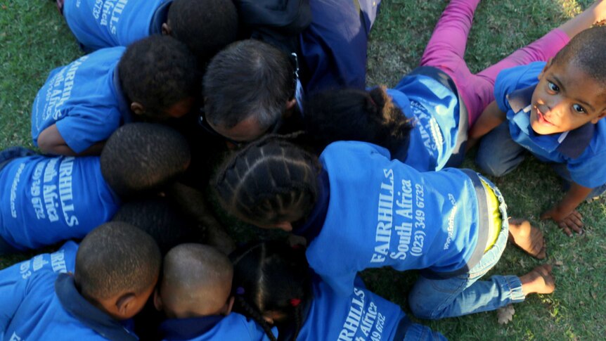 A group of African children in blue shirts bend over an iPad outside.