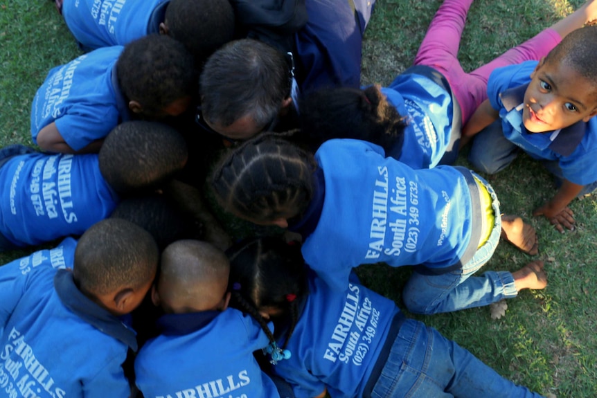 A group of African children in blue shirts bend over to look at an iPad outside.