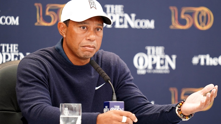 Tiger Woods holds out his hand while sitting in front of a sponsor board